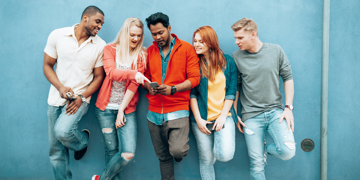 Group of GenZ young adults using a smartphone together, casually dressed, against a blue wall.